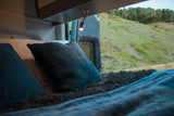 Cozy **rv rental** interior: a comfy bed with pillows, reading light, and a scenic view of a green hillside via open rear doors.