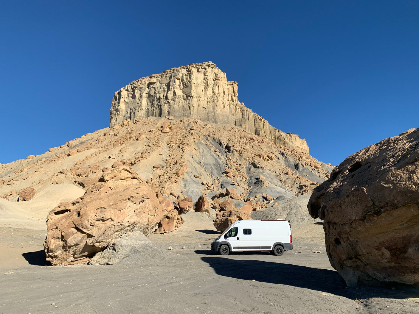 An RV rental van sits in a remote, rocky desert landscape, dwarfed by a large mesa under a clear blue sky.