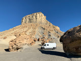 An RV rental van sits in a remote, rocky desert landscape, dwarfed by a large mesa under a clear blue sky.