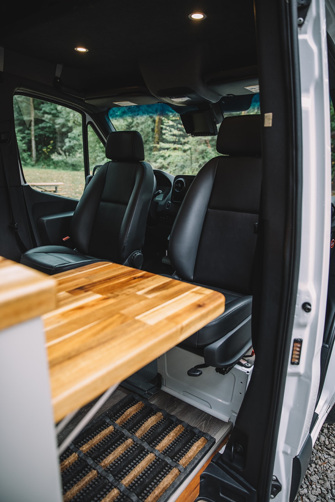  parked at a wooded campsite. Perfect for RV rental adventures.|A peek inside a sleek rv rental camper van: comfortable front cabin seats and a stylish custom-built living area with wood accents.|RV rental's cozy interior: kitchen with induction cooktop — photo 8