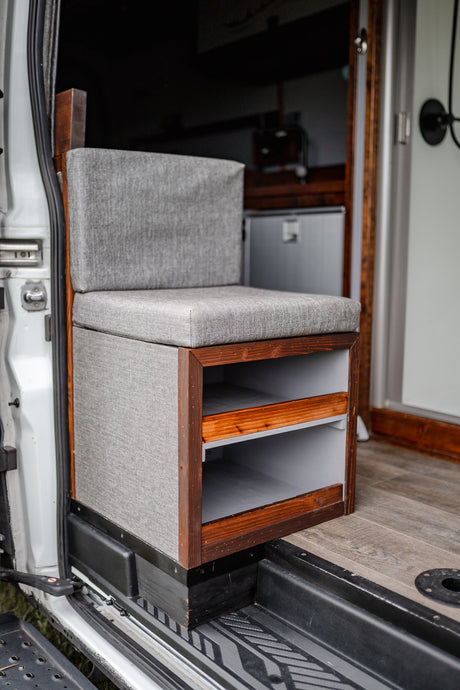 Cozy RV rental interior: grey upholstered seat with custom wood storage cubbies & integrated fridge for practical camper van living.