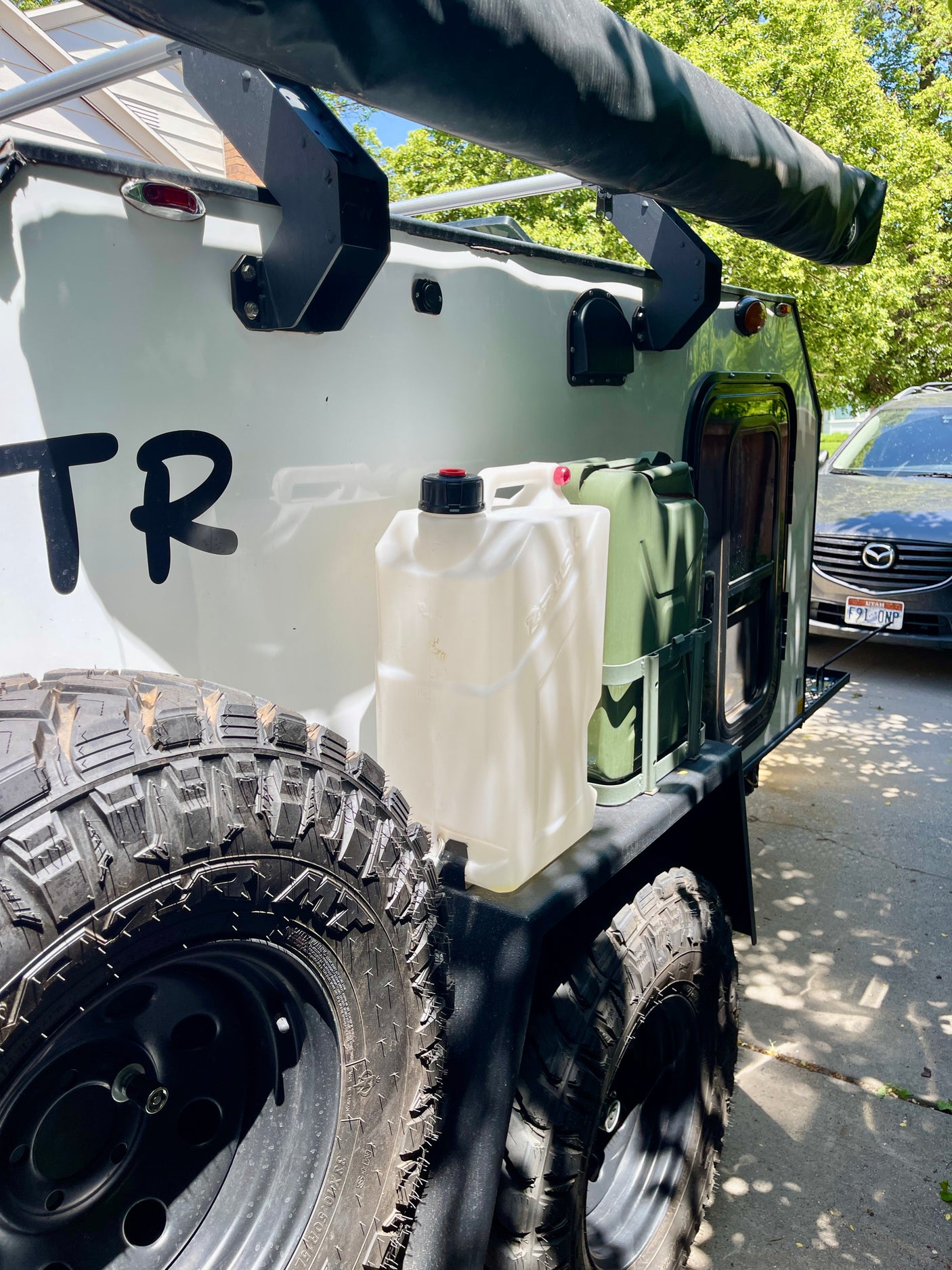Side view of a white off-road camper trailer, equipped with rugged tires, water/fuel containers, and an awning. Great for an rv rental.