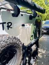 Side view of a white off-road camper trailer, equipped with rugged tires, water/fuel containers, and an awning. Great for an rv rental.