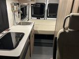 View inside an RV rental, featuring a compact kitchen with sink and stovetop, leading to a comfortable bed area with covered windows.