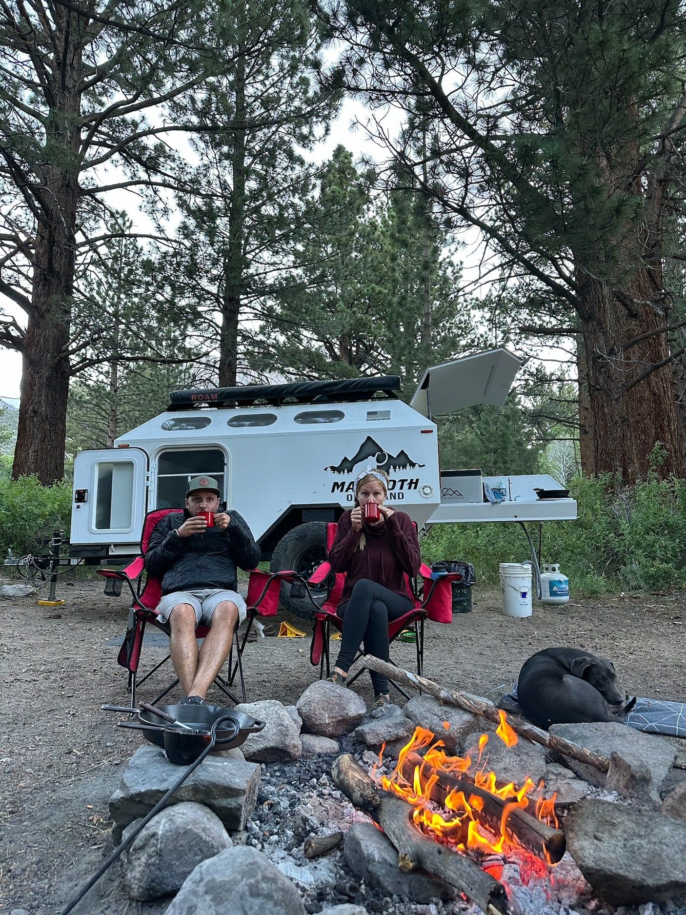 RV rental adventure! Two campers and their dog enjoy a cozy campfire by their off-road trailer under tall trees.