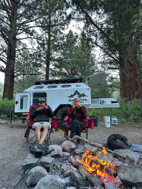 RV rental adventure! Two campers and their dog enjoy a cozy campfire by their off-road trailer under tall trees.
