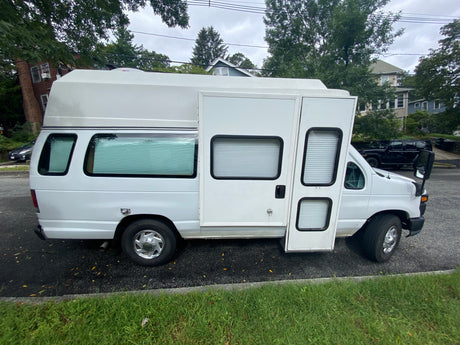 White Ford E-Series RV rental van, converted into a camper with high roof, side entry, and window blinds. Parked roadside.