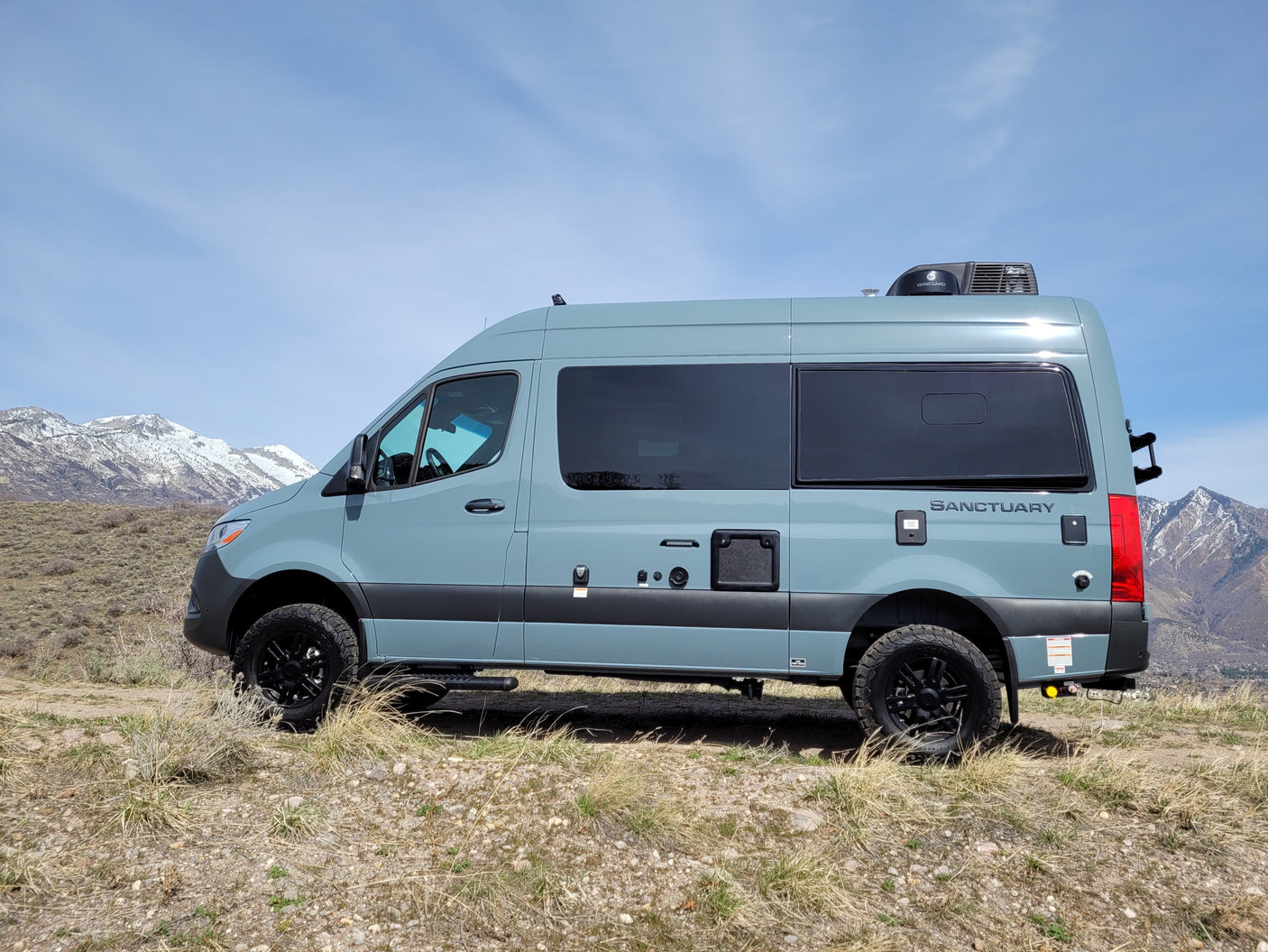 Explore with this light blue Sanctuary RV rental van, parked on a hill with impressive snow-capped mountains in the background.