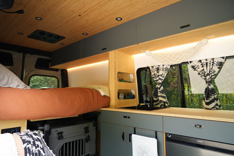 A modern RV rental van interior features a raised bed, compact kitchen, overhead storage, and nature views from the window.
