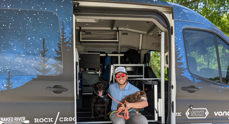 A man and his two dogs pose inside a spacious RV rental van with custom starry night wrap, ideal for outdoor trips.