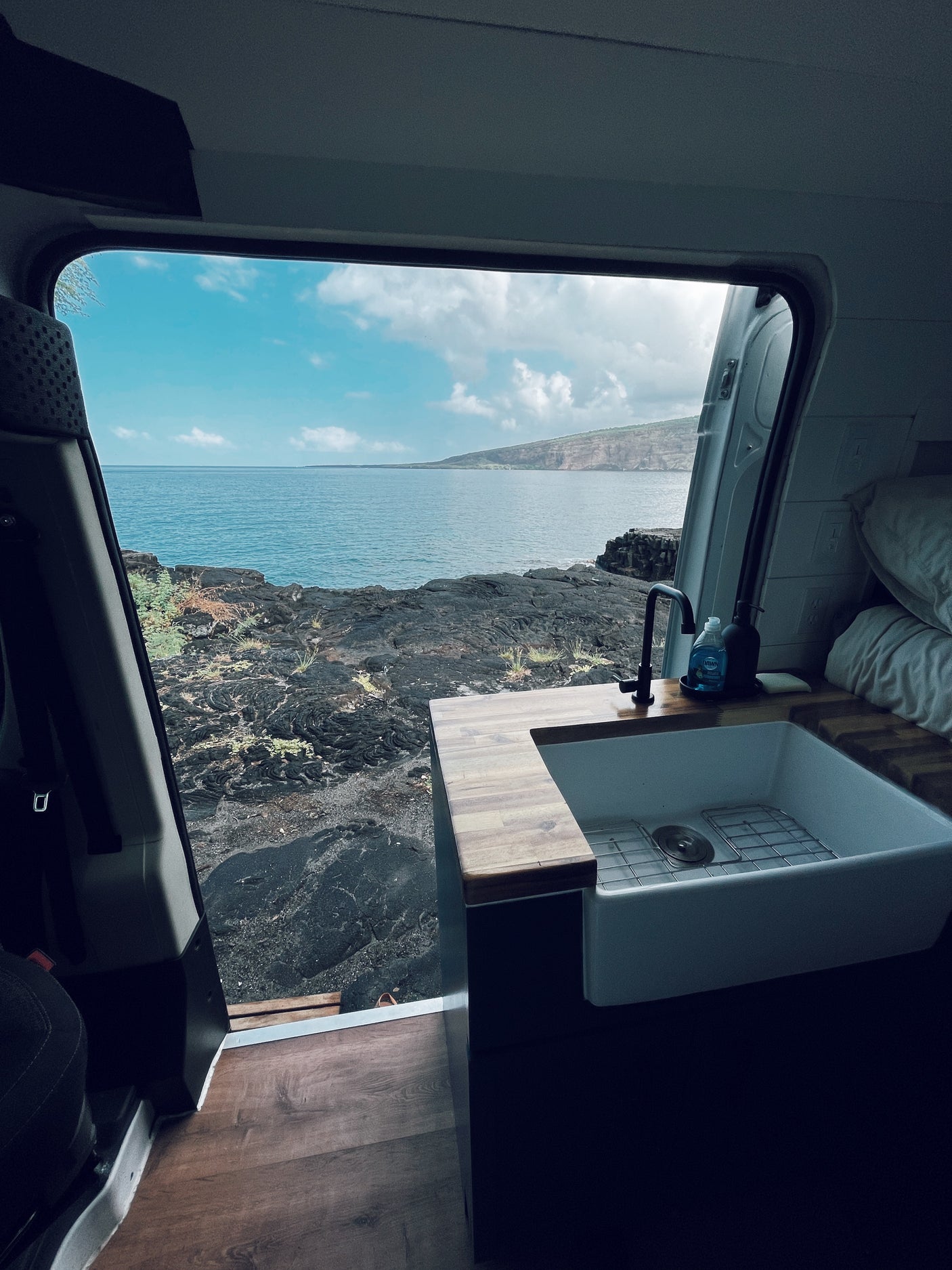 Inside an RV rental: a modern kitchen with a sink, facing a breathtaking view of the ocean and volcanic coastline. Ideal for van life.