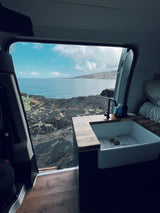 Inside an RV rental: a modern kitchen with a sink, facing a breathtaking view of the ocean and volcanic coastline. Ideal for van life.