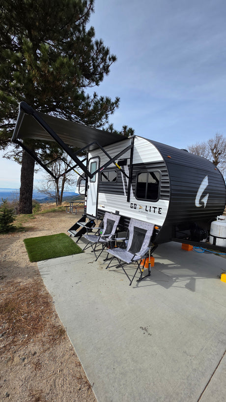 An rv rental, a Go Lite travel trailer, is set up with an awning and chairs on a concrete pad, overlooking a scenic mountain vista.