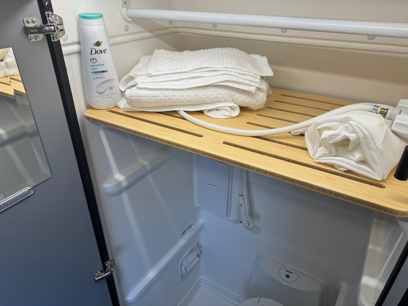 Compact RV rental bathroom, featuring a shower, toilet, fresh towels, and Dove body wash on a slatted wooden shelf.