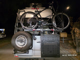 RV rental: A silver Mercedes van's rear shows a bike, spare tire, & side storage. Night scene, ready for adventure.