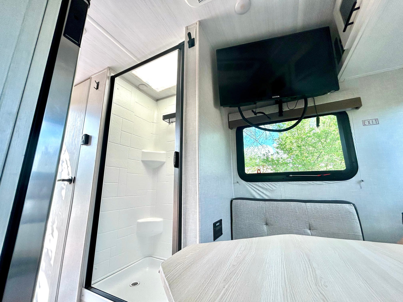 Bright RV rental interior featuring a walk-in shower, wall-mounted TV, dinette seating, and an exit window. — photo 9