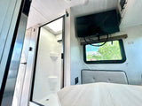Bright RV rental interior featuring a walk-in shower, wall-mounted TV, dinette seating, and an exit window.