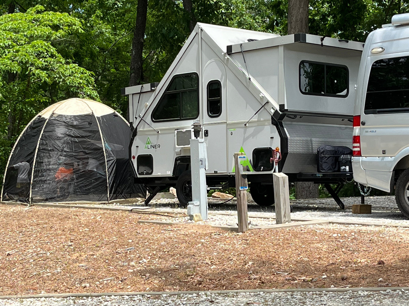 A white A-frame pop-up camper and RV van are parked at a wooded campsite with a screen tent, perfect for an rv rental.