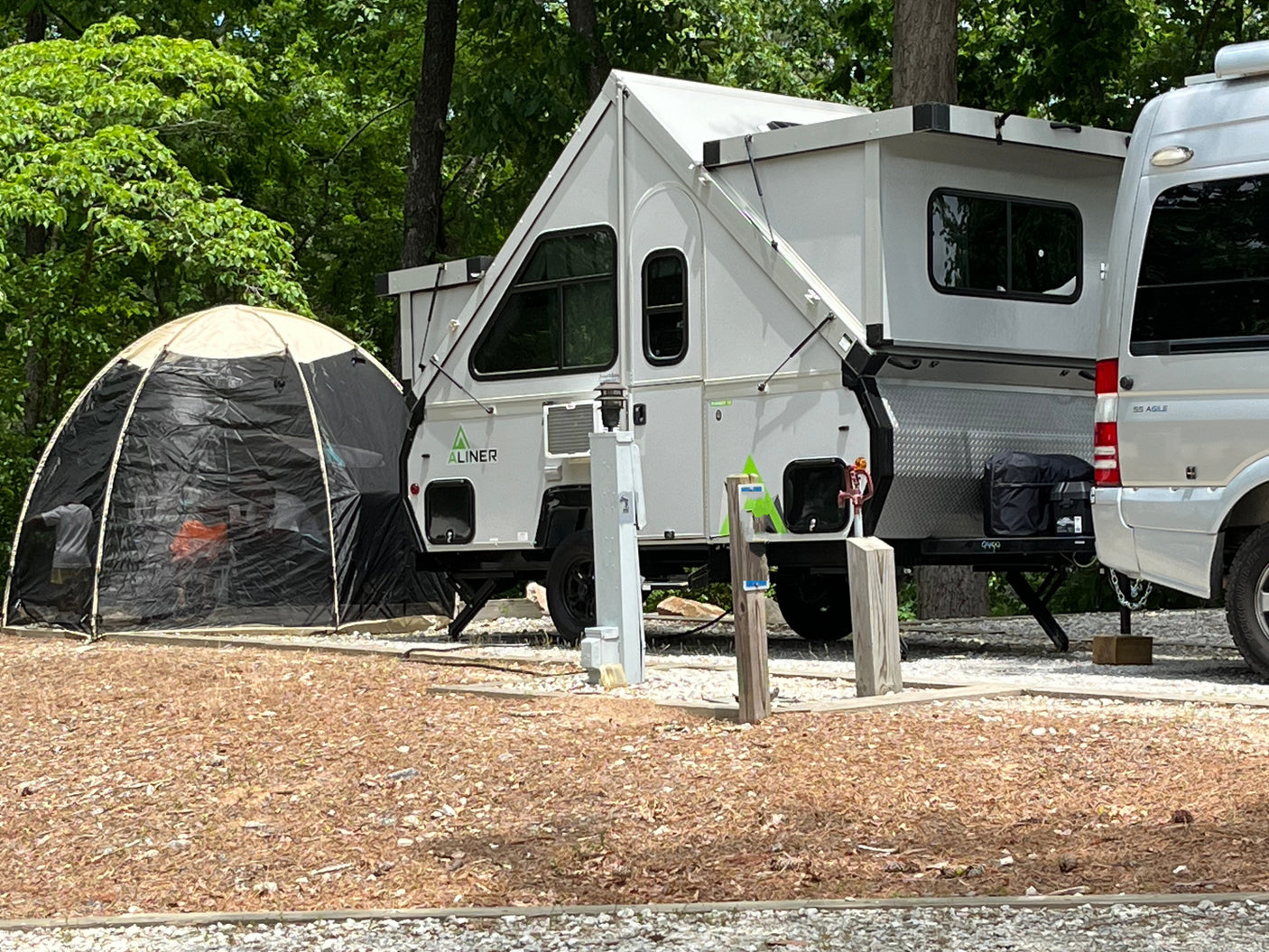 A white A-frame pop-up camper and RV van are parked at a wooded campsite with a screen tent, perfect for an rv rental.