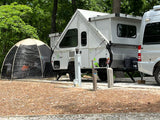 A white A-frame pop-up camper and RV van are parked at a wooded campsite with a screen tent, perfect for an rv rental.