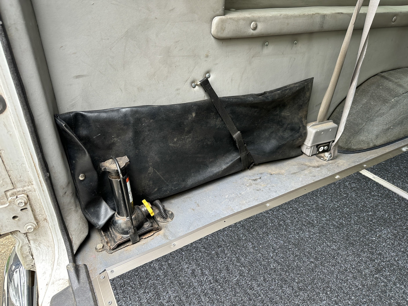 RV rental utility zone: jack, secured black storage bag, and seatbelt in a vehicle interior with a textured floor mat.