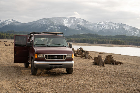 A maroon Ford RV rental van on a gravel lake shore with tree stumps and snow-capped mountains under an overcast sky.