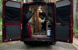 Rear view of a red RV rental with open doors, revealing a custom-built interior with bed platform & storage, parked in a forest.