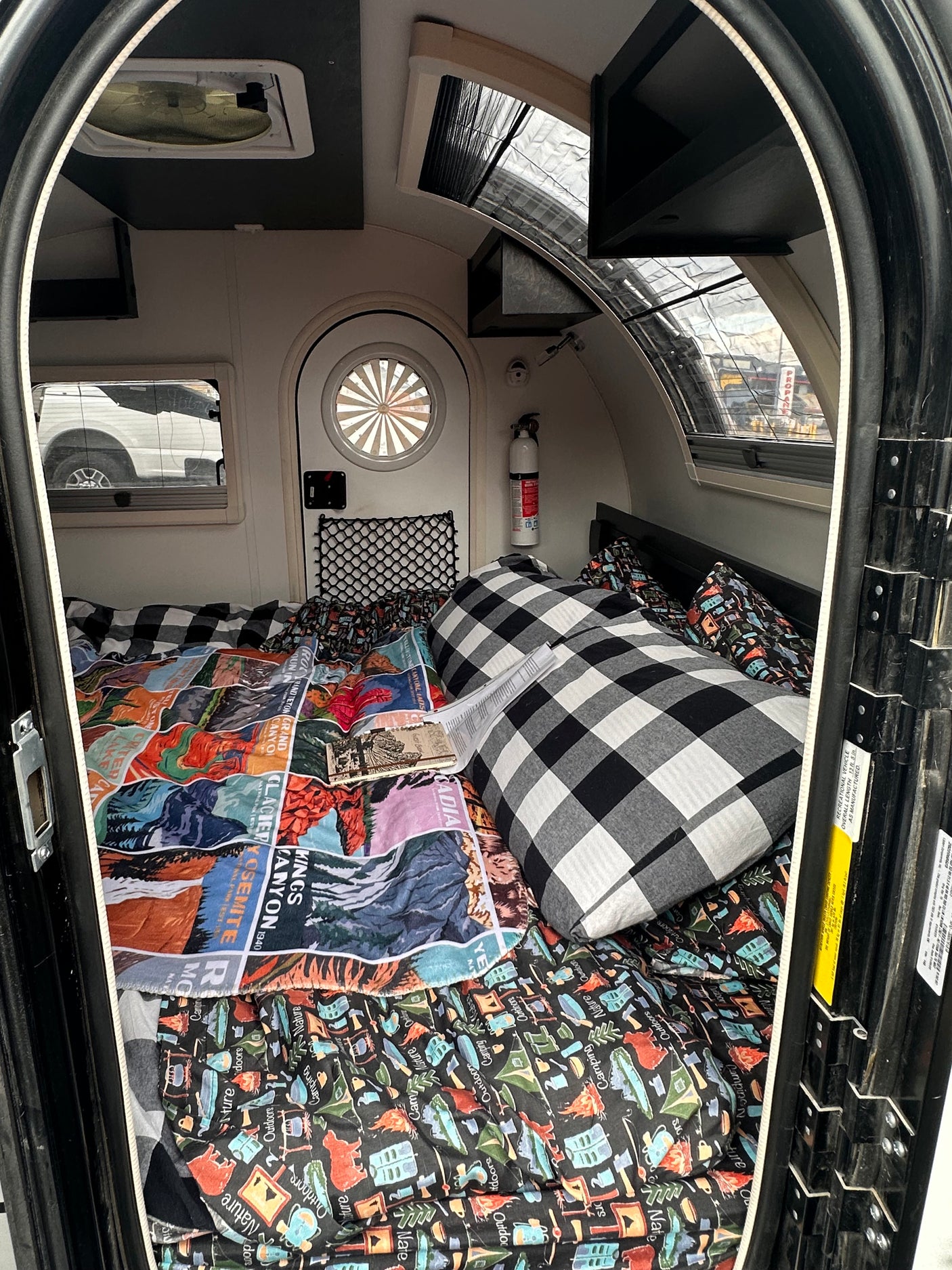 Inside a compact RV rental, featuring a cozy bed with patterned blankets, windows, and an overhead fan.