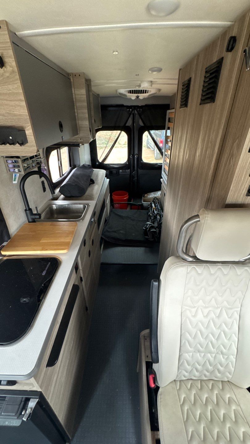 Compact RV rental interior featuring a kitchen with sink & stove, narrow aisle, and passenger seat. Ready for adventure!