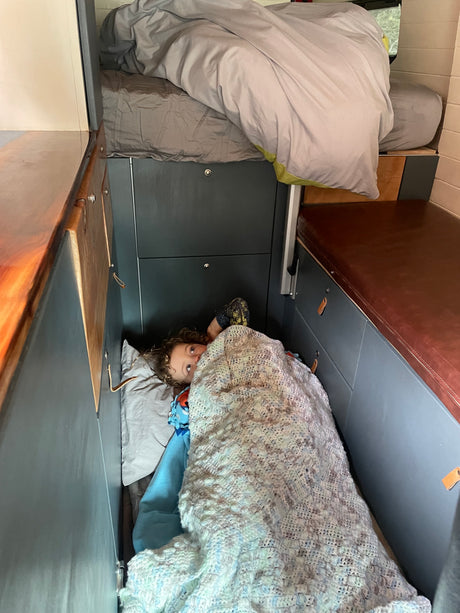 Cozy rv rental bunk: A child peeks from under a blanket in a compact camper van sleeping area, with an upper bunk visible.