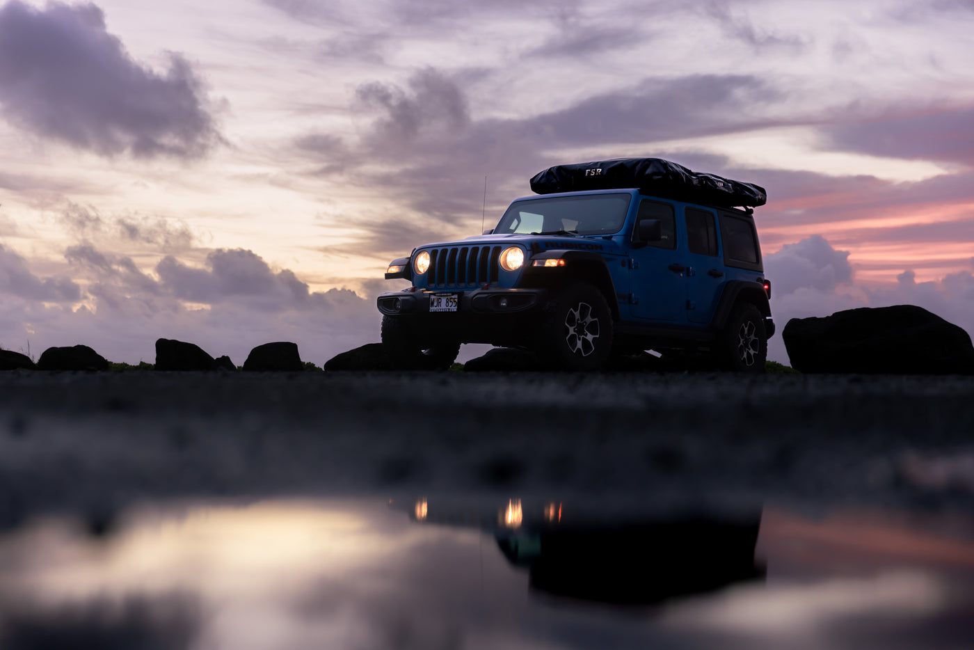 A blue Jeep with a roof tent is silhouetted against a colorful sunset sky, headlights reflecting in a foreground puddle. Ideal for an RV rental adventure. — photo 8