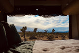 RV rental view: Cozy bed overlooking Joshua trees, desert landscape, and snowy mountains under a cloudy sky.