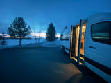 Experience winter travel with an RV rental! A white camper van glows from within at dusk, surrounded by snowy fields and trees.