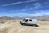 White RV rental van overlooks a desert landscape with mountains under a bright blue sky. Perfect for off-grid adventures.