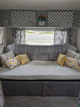Cozy RV rental interior: daybed with yellow & striped pillows, patterned blanket, checkered decor, and roadside window view.