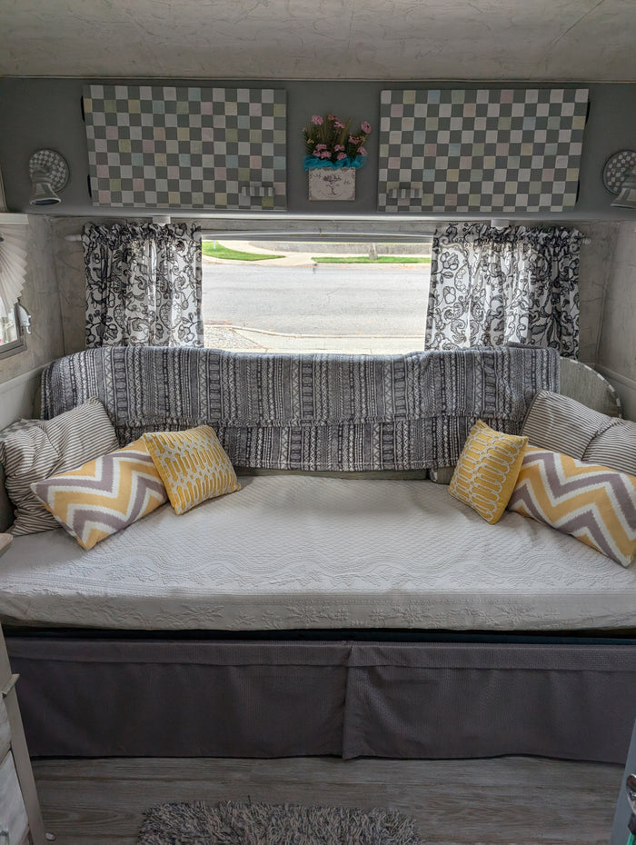 Cozy RV rental interior: daybed with yellow & striped pillows, patterned blanket, checkered decor, and roadside window view.