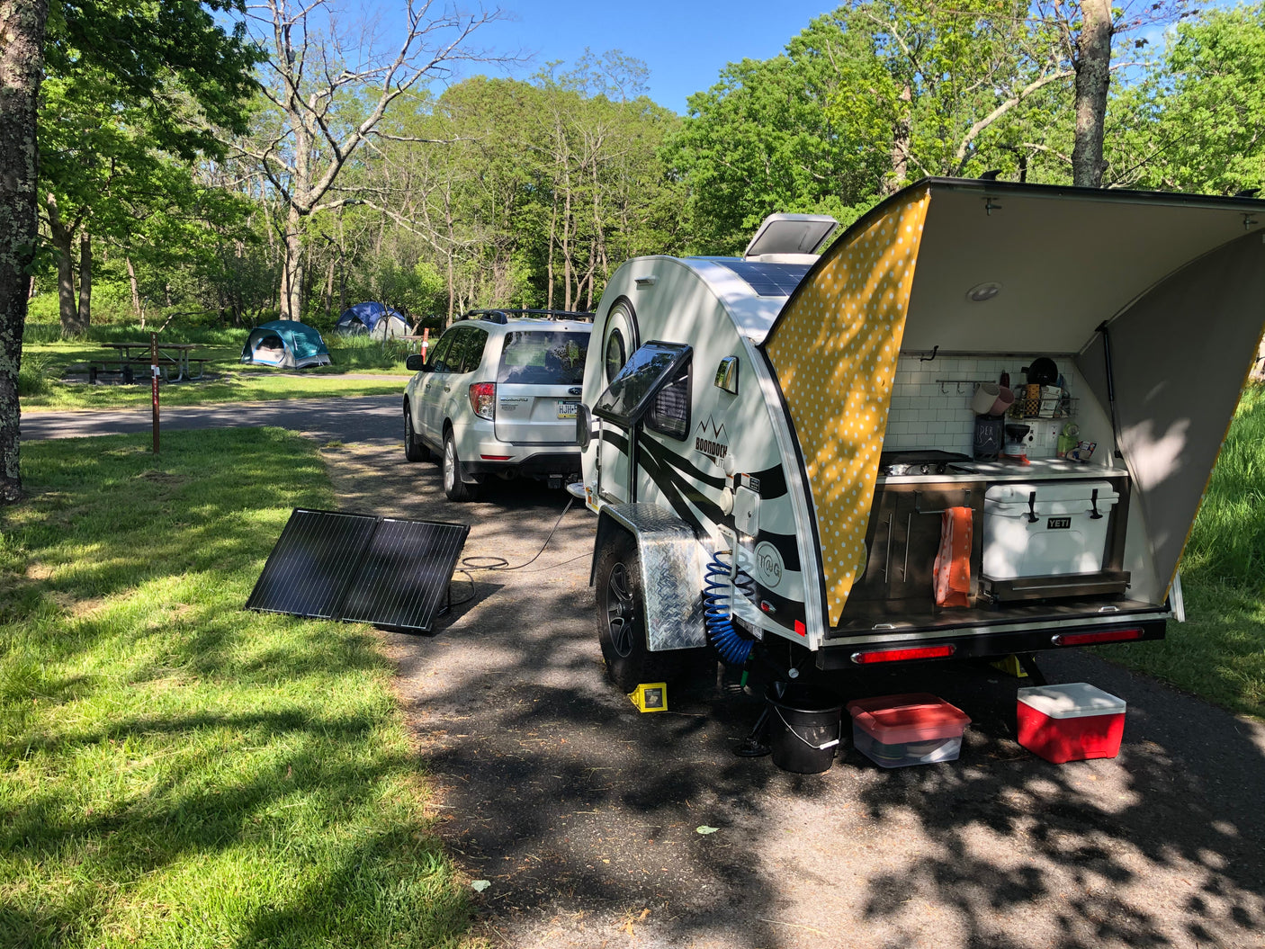 Teardrop RV rental at a sunny campsite, showcasing its open kitchen, solar panels, and connected SUV for outdoor trips.