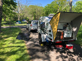 Teardrop RV rental at a sunny campsite, showcasing its open kitchen, solar panels, and connected SUV for outdoor trips.