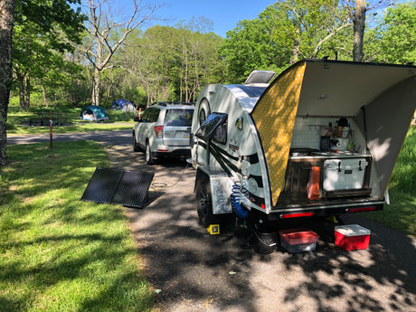 Teardrop RV rental at a sunny campsite, showcasing its open kitchen, solar panels, and connected SUV for outdoor trips.