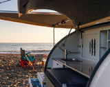Experience serene beach camping with an RV rental! This setup shows an open teardrop trailer kitchen by the ocean.