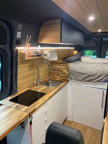 RV rental interior: modern kitchen with wood counters, induction cooktop, sink, white cabinets, and a comfortable bed.