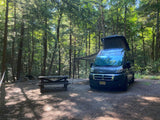 An RV rental camper van with a pop-top and awning set up at a forest campsite. A picnic table and fire pit complete the outdoor scene.