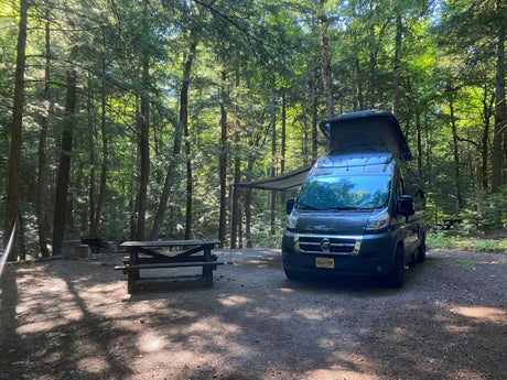 An RV rental camper van with a pop-top and awning set up at a forest campsite. A picnic table and fire pit complete the outdoor scene.