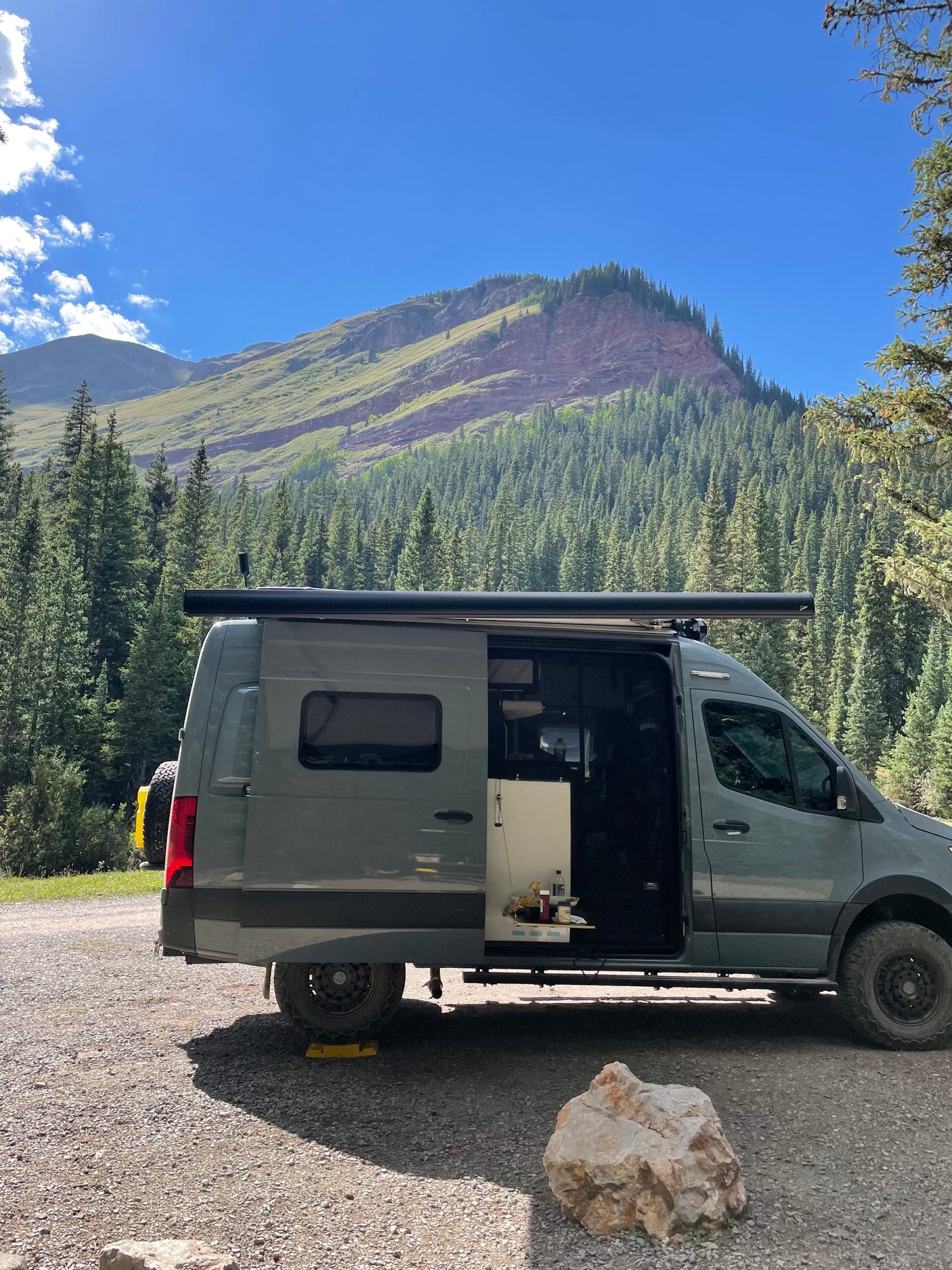 RV rental camper van with open door on gravel, showing interior. Majestic mountains and dense forest in background. Perfect for nature trips.