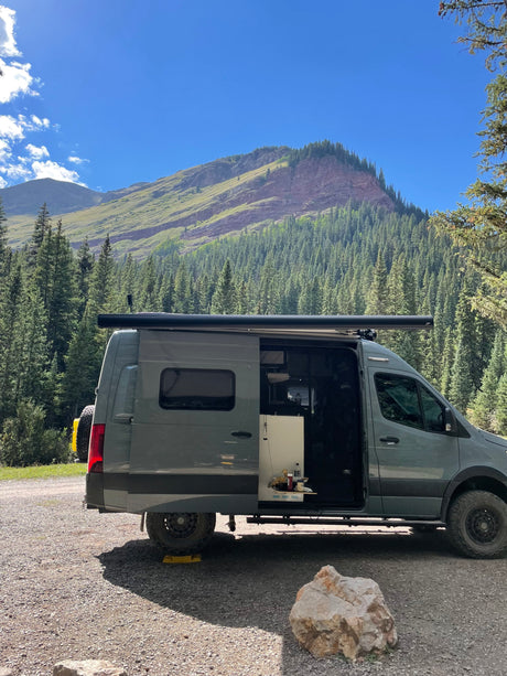 RV rental camper van with open door on gravel, showing interior. Majestic mountains and dense forest in background. Perfect for nature trips.