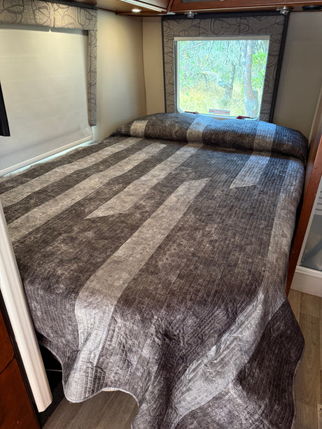 RV rental bedroom: a grey geometric-quilted comforter on the bed, with a window showing trees. Comfortable travel accommodation.