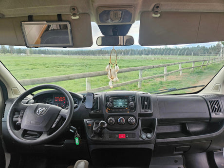 Dashboard view inside an rv rental with Dodge steering wheel, tablet, dream catcher, overlooking a green field and fence.