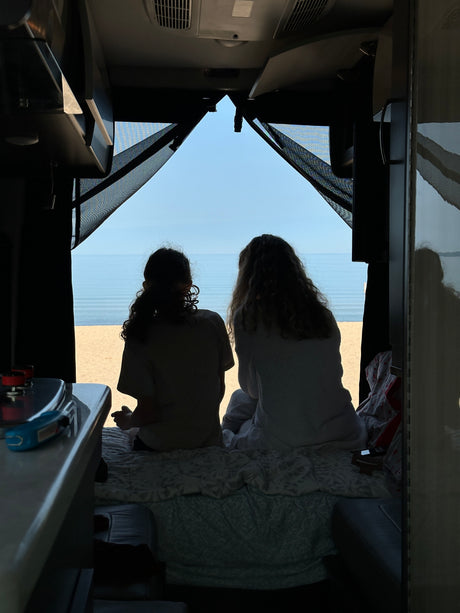 Two people enjoy a serene beach view from their RV rental. Inside, silhouetted figures gaze at the ocean and sandy shore.