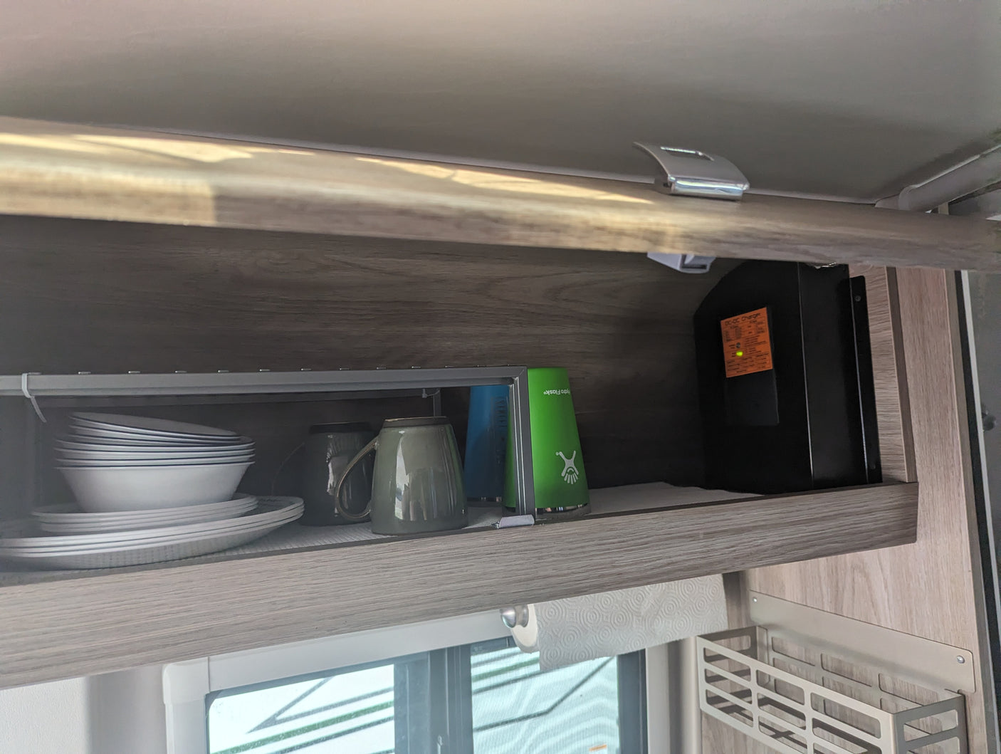 An RV rental's overhead cabinet neatly stores dishes, mugs, water bottles, and a DC-DC charger, with paper towels below.