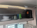 An RV rental's overhead cabinet neatly stores dishes, mugs, water bottles, and a DC-DC charger, with paper towels below.